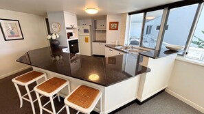 A modern kitchen with a large island, white cabinetry, and a window providing a view. - Ocean Shores Apartment 13 (SOUTH WEST ROCKS)