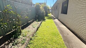 A narrow pathway flanked by a brick building and a metal fence, leading to a grassy area. - Beach House - South West Rocks (SOUTH WEST ROCKS)