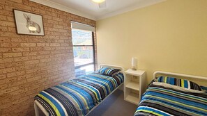 A room with two beds, a brick wall, a window, and a picture of a rabbit. - Beach House - South West Rocks (SOUTH WEST ROCKS)
