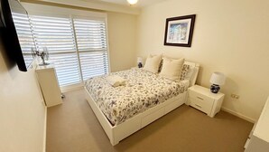 A bedroom with a bed, bedside tables, a lamp, a picture on the wall, and a window with blinds. - Jubilee Apartment 14 (South West Rocks)