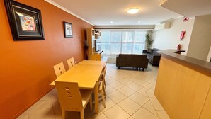 A dining area with a wooden table and chairs, a kitchen area with a counter, and a living room with a sofa. - Jubilee Apartment 14 (South West Rocks)