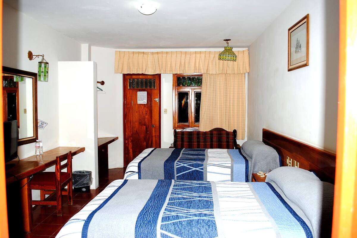 Une chambre d’hôtel avec deux lits, un bureau, une chaise, une poubelle et une fenêtre avec des rideaux.