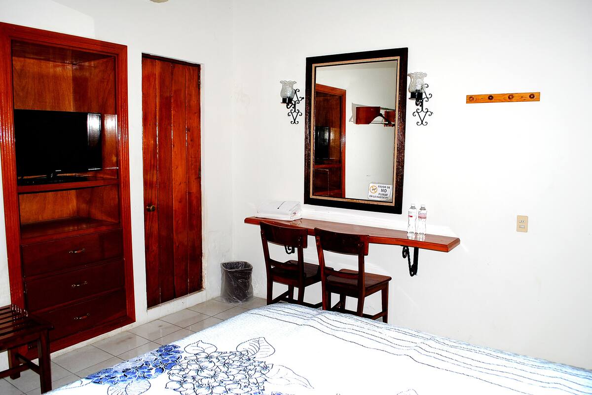 Une chambre avec une armoire en bois, un bureau doté d’un miroir et un lit recouvert d’une couvre-lit à motifs floraux.