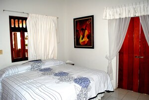 A bedroom with a bed, a window with curtains, a framed picture on the wall, and a red door.