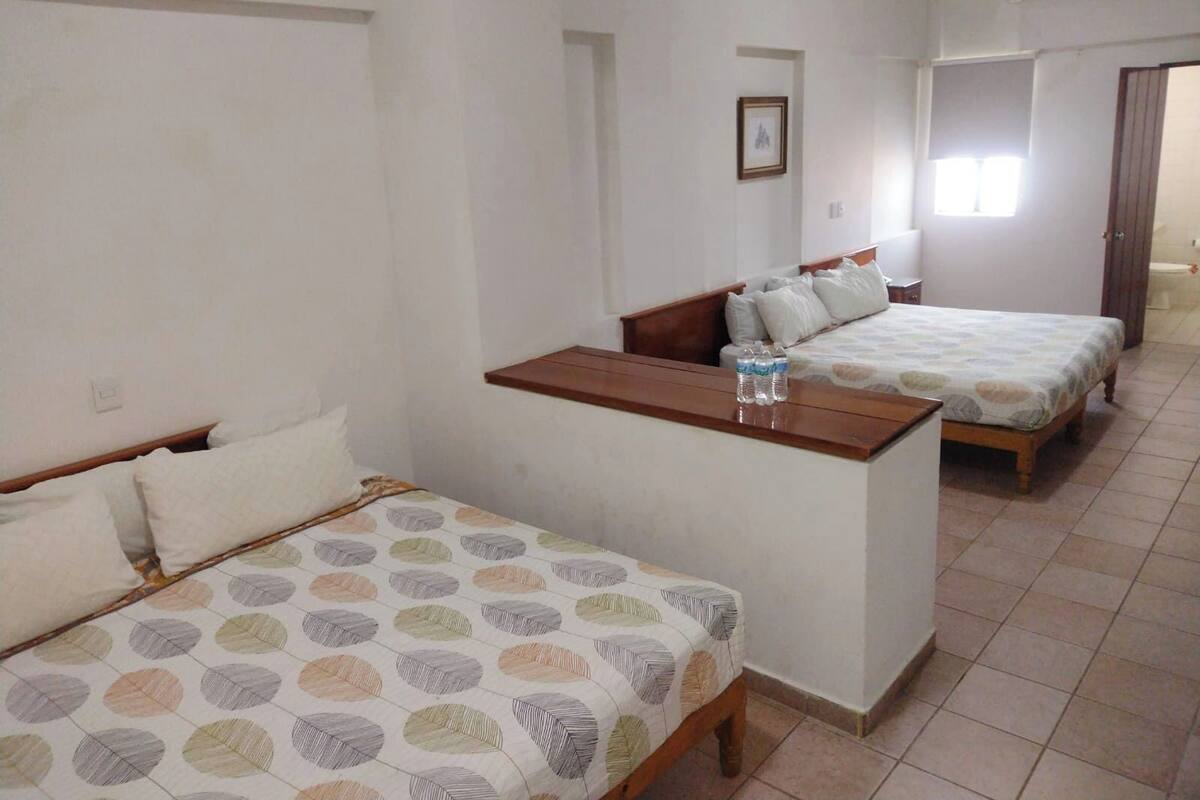 Une chambre avec deux lits simples, une petite table en bois avec des bouteilles d’eau et un tableau encadré au mur.