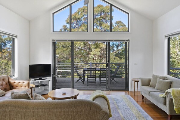 A modern living room with a large TV, comfortable seating, a dining area, and a balcony with a view of trees.