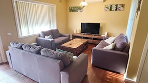 A living room with a sofa set, a coffee table, a TV, and a window with blinds. - Seamist Cottage - Pet Friendly (SOUTH WEST ROCKS)