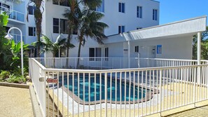 A pool area with a white fence, a sign indicating a garage door, and a building with multiple windows and balconies in the background. - Ocean Shores Apartment 17 (SOUTH WEST ROCKS)