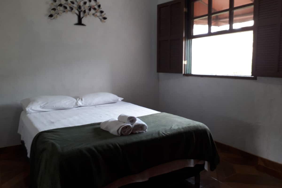 A room with a bed, a window with brown shutters, and a wall decoration of a tree.