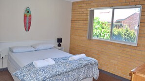 A bedroom with a bed, pillows, a nightstand, a lamp, a surfboard on the wall, and a window with a view of greenery and a building. - Wards Holiday Flat 7 (SOUTH WEST ROCKS)