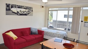 A room with a red sofa, a bed with a patterned bedspread, a wooden table with a bowl, and a window with a view of buildings. - Wards Holiday Flat 7 (SOUTH WEST ROCKS)