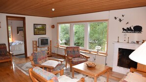 Una sala de estar acogedora con muebles de madera, chimenea y ventanas amplias que dan a un paisaje de árboles.