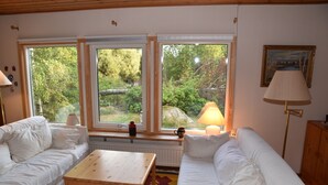 Una habitación con dos sofás blancos, una mesa de madera y una alfombra colorida. Se ven amplias ventanas con vistas a zonas verdes.