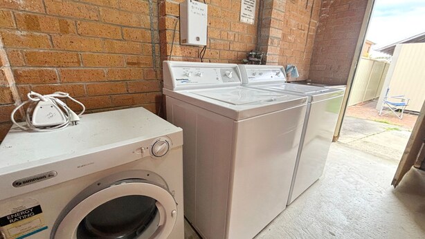A laundry area with a front-loading washer and dryer, a power outlet, and a brick wall. - Clarkes Flat 4 - walk to the beach (South West Rocks)