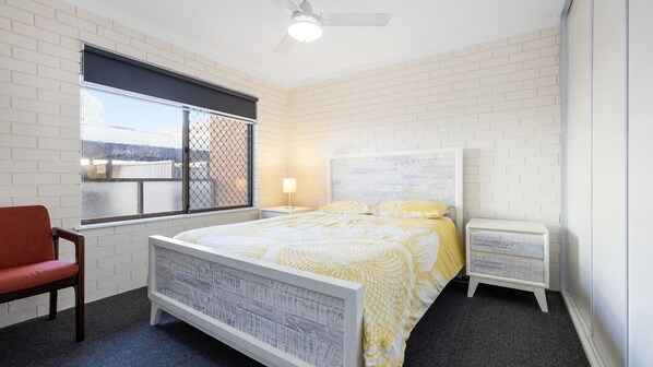 Ein Schlafzimmer mit weißer Ziegelwand, einem Bett mit gelb-weißer Tagesdecke, einem roten Sessel, einem Nachttisch mit Lampe und einem Deckenventilator.