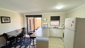 A compact kitchen with a dining area, white brick walls, a microwave, a refrigerator, and a window with a view. - Cameo Court 1 - walk to the beach - Linen Supplied (SOUTH WEST ROCKS)