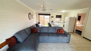 A living room with a grey sectional sofa, a ceiling fan, and a kitchen area with a refrigerator and microwave. - Cameo Court 1 - walk to the beach (SOUTH WEST ROCKS)