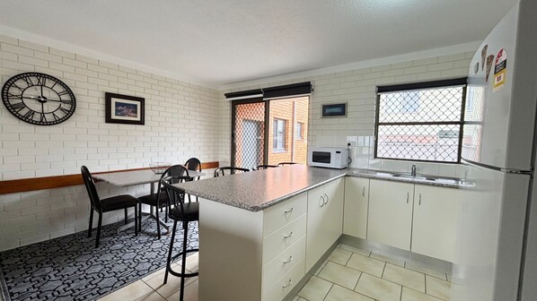 A kitchen with white cabinets, a microwave, a refrigerator, a clock, and a dining area with chairs and a table. - Cameo Court 1 - walk to the beach - Linen Supplied (SOUTH WEST ROCKS)