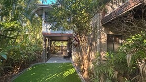 A covered outdoor area with a pathway leading to a building, surrounded by lush greenery.