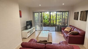 A living room with a TV, leather sofa, and a glass coffee table. - Amaroo - walk to the bay - Bring your own linen (ARAKOON)