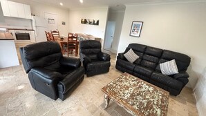 A living room with a black leather sofa, a matching armchair, a coffee table, and a dining area with a wooden table and chairs. - Three/Fifty Four Straight St Hat Head - Bring your own linen (Hat Head)