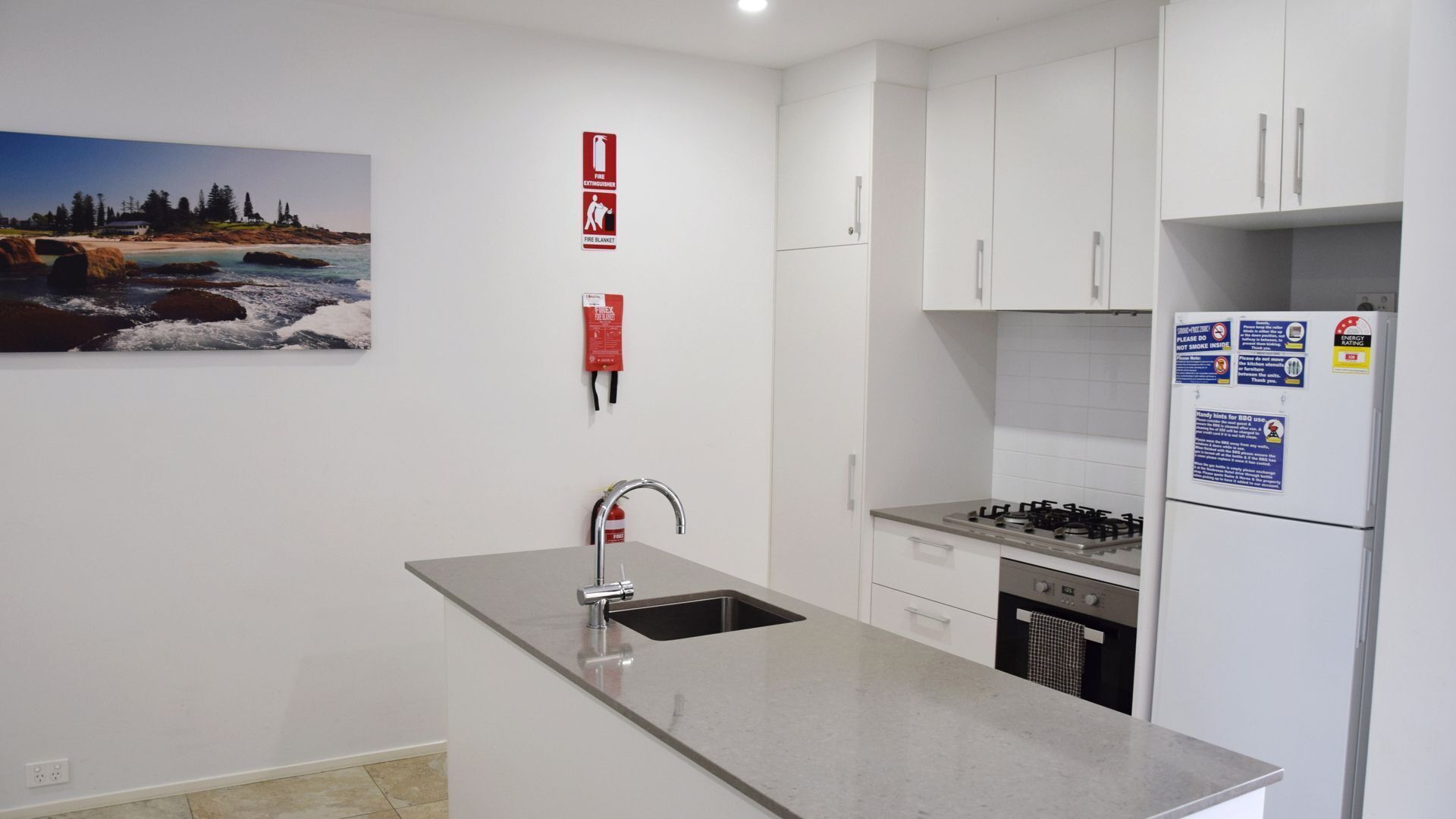 A modern kitchen with white cabinets, a stainless steel sink, a gas stove, and a refrigerator. There are fire safety signs on the wall and a painting of a beach scene.
