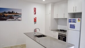 A modern kitchen with white cabinets, a stainless steel sink, a gas stove, and a refrigerator. There are fire safety signs on the wall and a painting of a beach scene. - Waterford Apartment 7 (SOUTH WEST ROCKS)