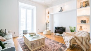 A modern living room with a sofa, coffee table, and a television. There is a bookshelf with decorative items and a large window providing natural light. - Estrela Neoclassical Apt River View (Lisbon)