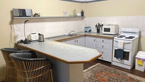 Een compacte keuken met een inbouwfornuis, magnetron en wastafel. Er staan twee barstoelen en een klein vloerkleed op de vloer.