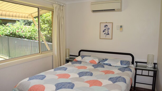A bedroom with a bed, a window, an air conditioner, a bedside table, and a framed picture on the wall. - Adaco's '19 On Sturt' - Bring your own linen (SOUTH WEST ROCKS)