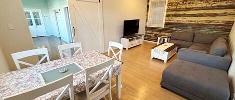 A living room with a sofa, a TV, a dining table with chairs, and a wooden floor.