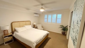 Une chambre à coucher comprenant un lit, des tables de chevet, un ventilateur de plafond, une fenêtre avec des stores et une plante en pot.