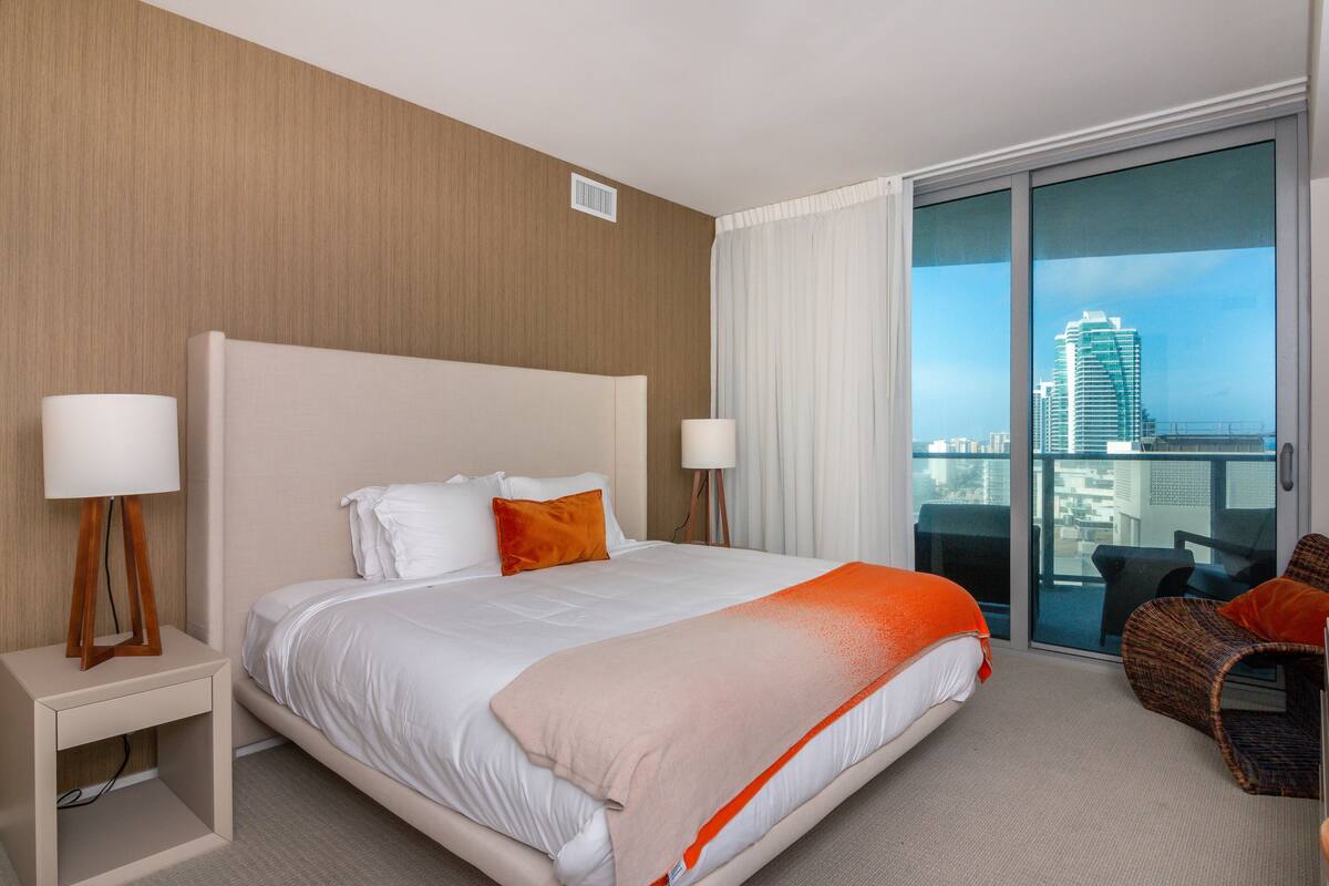 Ein Hotelzimmer mit einem großen Bett, zwei Nachttischen mit Lampen, einem Balkon mit Blick auf die Stadt und einem Korbstuhl.