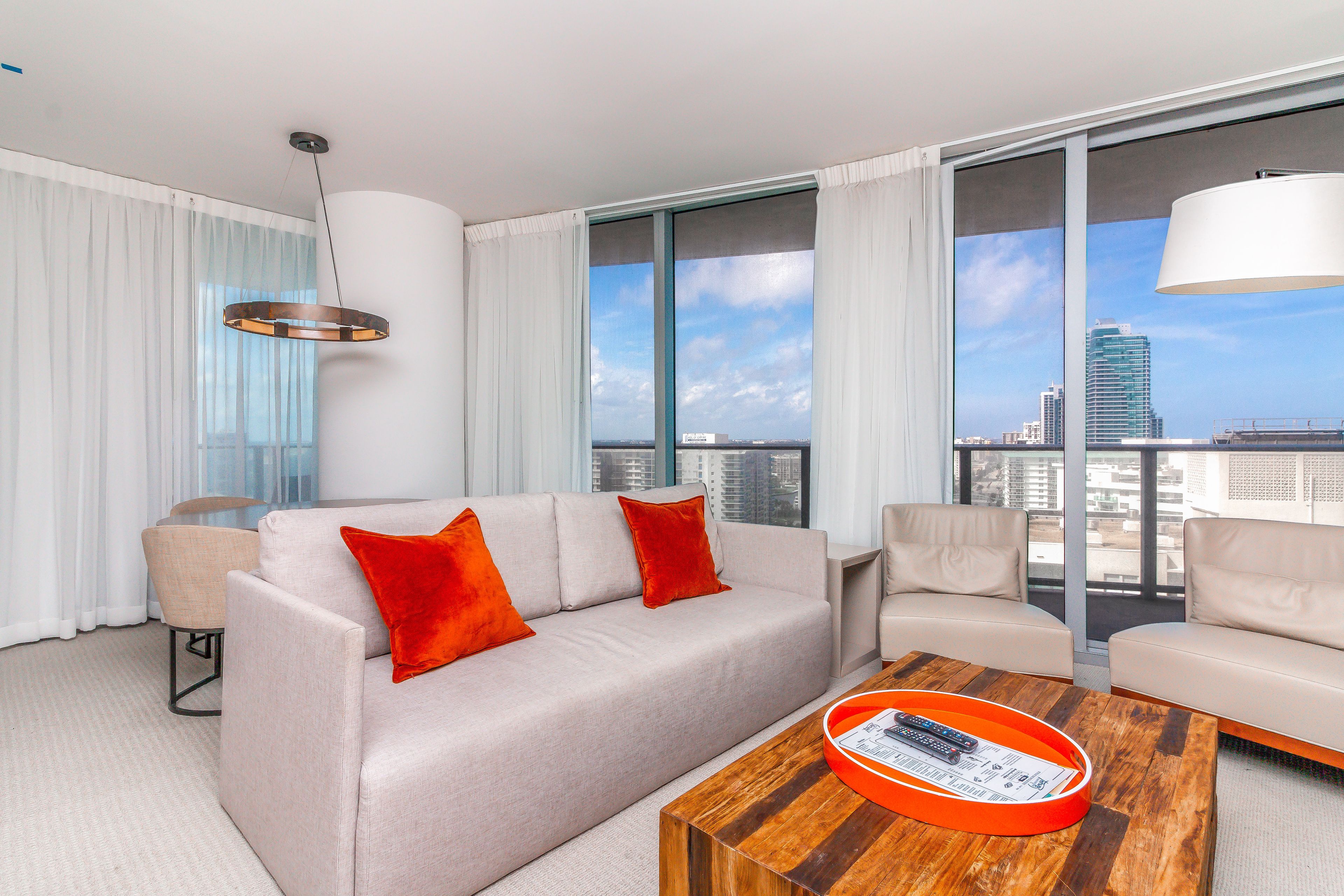 Ein modernes Hotelzimmer mit großem Fenster und Blick auf die Stadt, ein beiges Sofa mit roten Kissen, ein Holztisch und eine hängende Leuchte.
