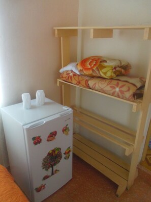 Quarto compacto com uma geladeira branca decorada com adesivos de frutas e legumes, uma pequena mesa branca com duas xícaras, e uma cama beliche de madeira com roupa de cama estampada.
