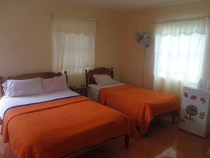 A room with two beds, each with an orange bedspread, a wooden headboard, and white pillows. There is a small table with a fan, and a refrigerator with fruit magnets. - Casa Yaquelin y Osmani (Viñales)