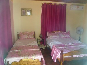 Quarto com duas camas, cada uma com roupa de cama rosa e estampas florais. Há uma pequena mesa com um abajur entre as camas, e um ar condicionado fixado na parede acima da cama à direita.