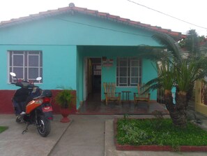 Um edifício de dois andares com telhado vermelho e paredes verde-água. Uma motocicleta está estacionada na frente, e há um pequeno jardim com uma palmeira.