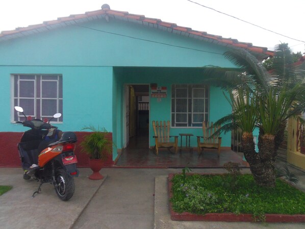 A two-story building with a red roof and teal walls. There is a motorcycle parked in front, and a small garden with a palm tree. - Casa Yaquelin y Osmani (Viñales)
