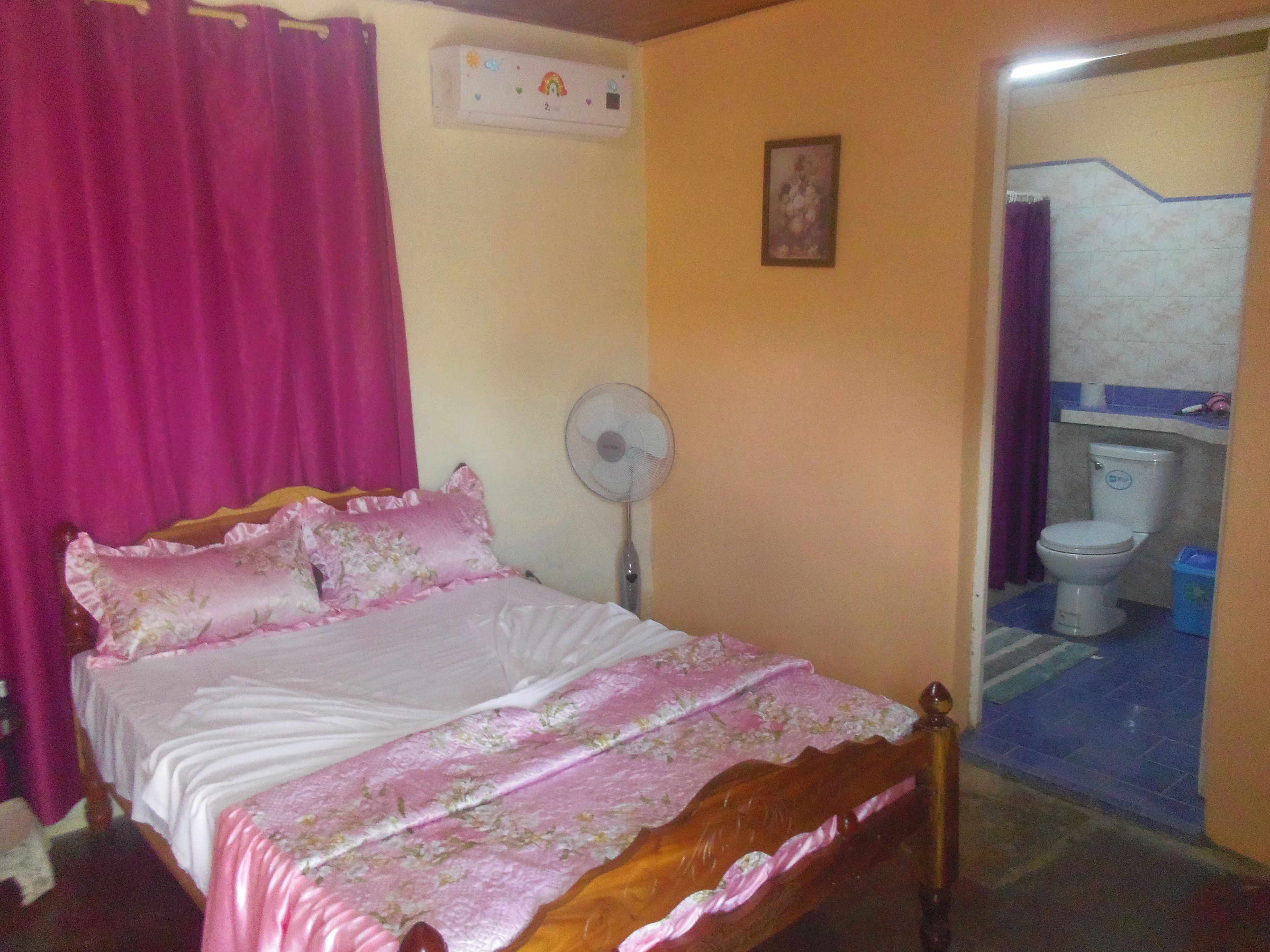 A bedroom with a bed, a fan, a bathroom visible through an open door, and a wall-mounted air conditioner.