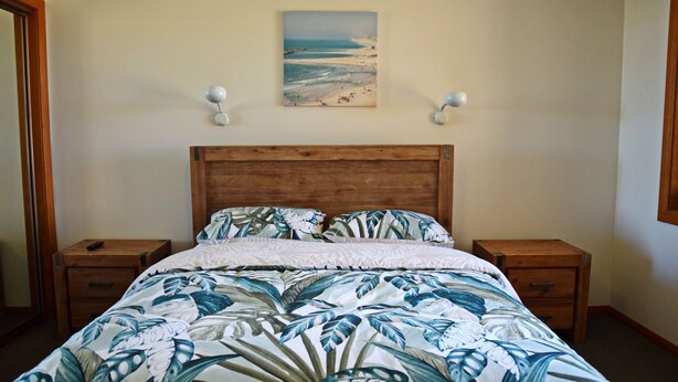 Ein Schlafzimmer mit einem hölzernen Kopfteil, einem Bett mit Bettdecke mit tropischem Blattmuster, zwei Nachttischen und einem Bild einer Strandlandschaft an der Wand.
