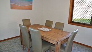 Ein Essbereich mit einem Holztisch und sechs Stühlen, ein Schachbrett auf dem Tisch, und ein Fenster mit einem Maschendrahtzaun davor.