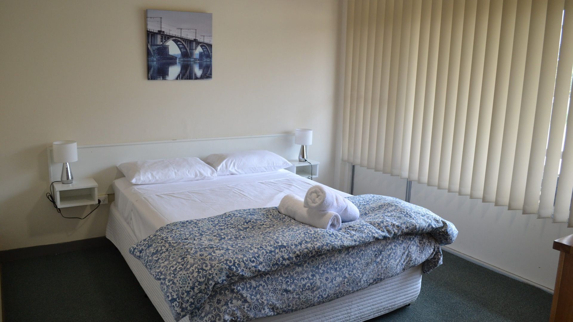 Une chambre d’hôtel avec un lit, du linge de lit blanc, une couverture à motifs, une serviette enroulée, une table de chevet avec une lampe et un tableau représentant un pont accroché au mur.