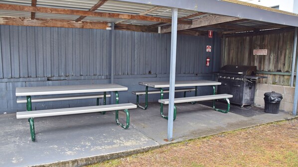 An outdoor covered picnic area with metal tables and benches, a barbecue grill, and a trash can. - Wards Holiday Flat 1 (SOUTH WEST ROCKS)