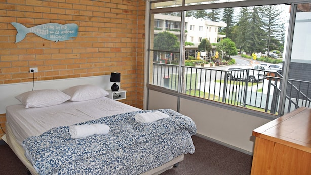 A bedroom with a bed, a nightstand, a lamp, and a window with a view of a building and street. - Wards Holiday Flat 8 (SOUTH WEST ROCKS)