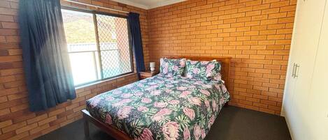 A bedroom with a brick wall, a bed with floral bedding, a window with curtains, and a ceiling fan.