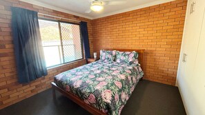 A bedroom with a brick wall, a bed with floral bedding, a window with curtains, and a ceiling fan. - Cameo Court 2 - walk to the beach (SOUTH WEST ROCKS)