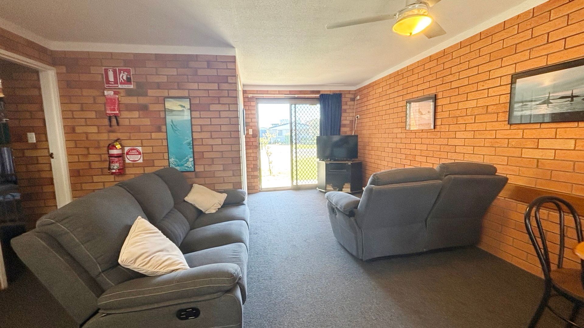 A living room with a brick wall, a sofa, a recliner, a television, and a door leading outside.