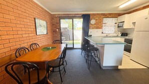 A compact kitchen with a dining area, featuring a brick wall, a wooden table, and a small island with stools. - Cameo Court 2 - Bring your own linen (SOUTH WEST ROCKS)