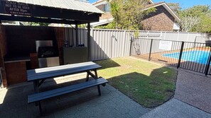 A barbecue area with a grill and seating, a fenced pool, and a grassy yard. - Cameo Court 2 - Bring your own linen (SOUTH WEST ROCKS)